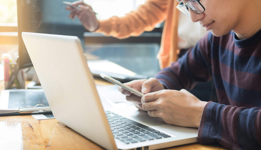 A graduate sitting in front of a laptop while holding a mobile phone